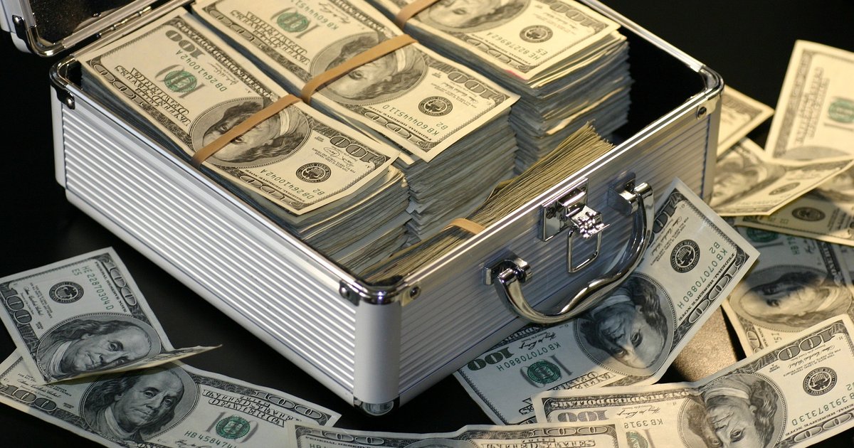 Open aluminum briefcase packed with banded stacks of hundred-dollar bills, with loose bills scattered around it