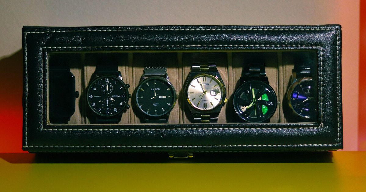 Six wristwatches in a black leatherette display box with a glass lid